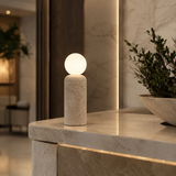 A modern table lamp with a cylindrical textured stone base and a glowing spherical shade, placed on a polished marble countertop in a stylish interior setting.