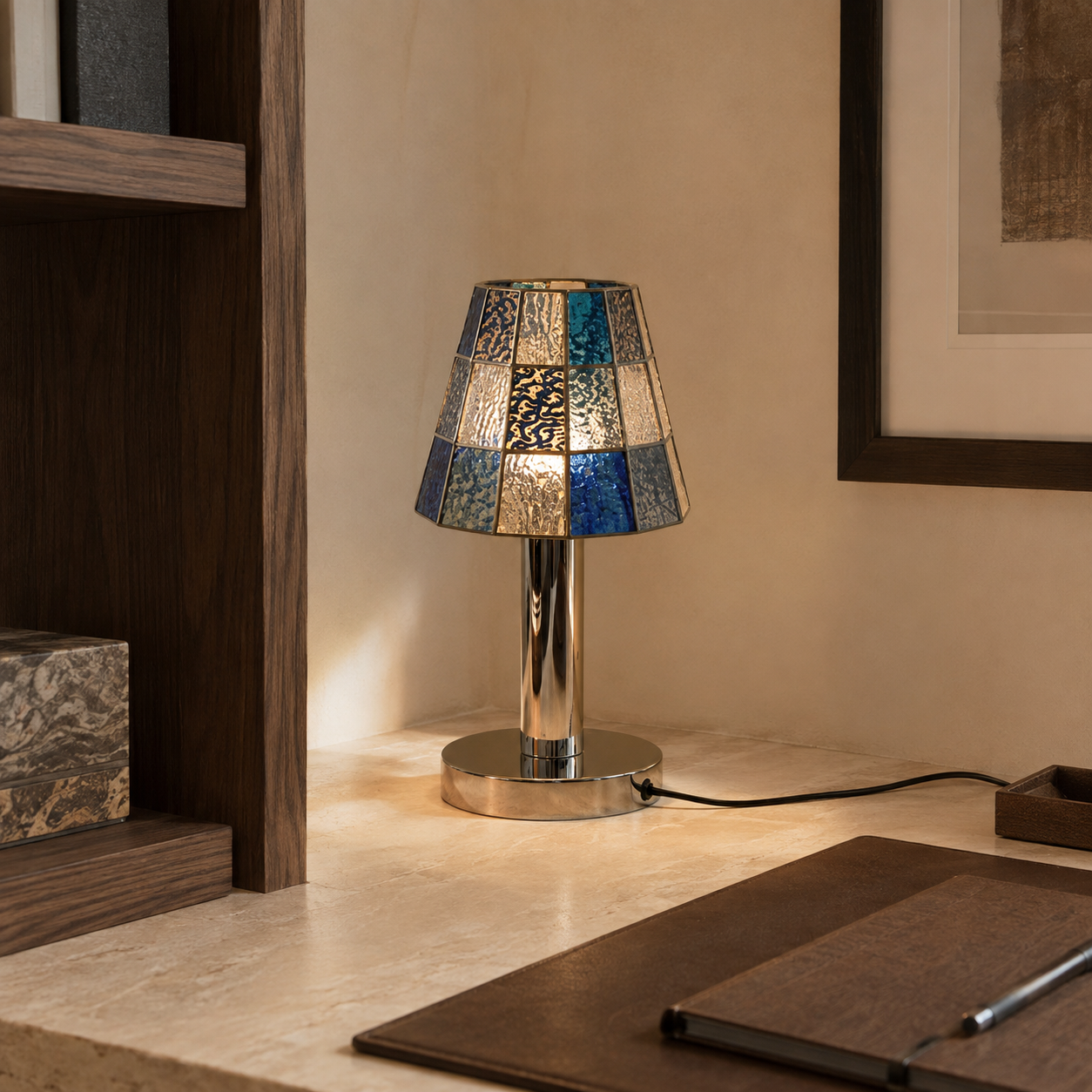 A stained glass table lamp with blue, silver, and gold patterned shades and a polished silver base, lit on a beige stone desk in a warm interior setting.