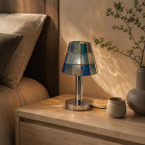 A table lamp with a mosaic blue and silver stained-glass shade and a polished silver base sits on a light wood bedside table in a warmly lit bedroom setting.