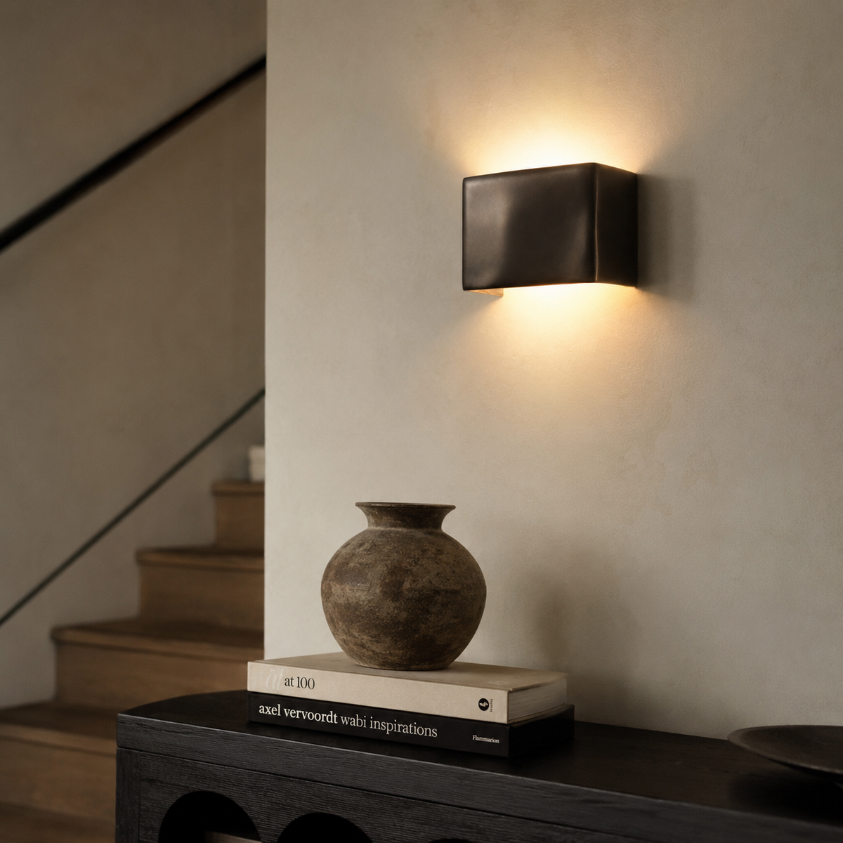 A modern rectangular wall sconce with a dark bronze finish, mounted on a textured beige wall and casting a warm light downward. Below, a dark wooden console table holds a ceramic vase on stacked books in a softly lit interior setting.