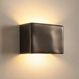 A modern square wall sconce light fixture with a dark bronze finish, mounted on a textured beige wall and casting a warm yellow light upward and downward.