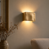 A modern square wall sconce with a textured metallic gold finish, mounted on a beige wall and casting a warm yellow light downward and upward. The scene is styled with a vase of dried branches on the left and the corner of a beige sofa at the bottom right.