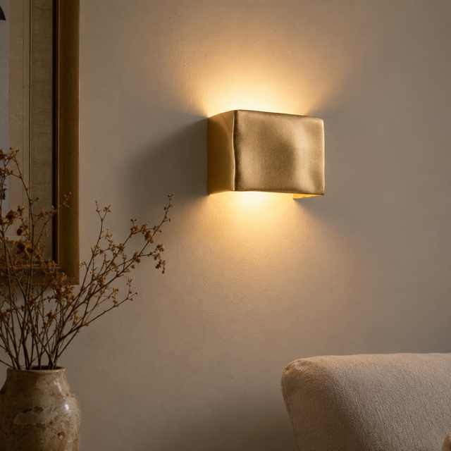 A modern square wall sconce with a textured metallic gold finish, mounted on a beige wall and casting a warm yellow light downward and upward. The scene is styled with a vase of dried branches on the left and the corner of a beige sofa at the bottom right.