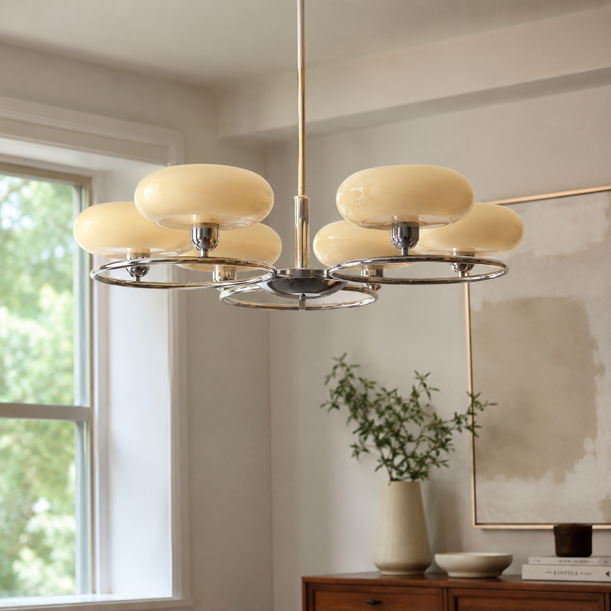 A modern six-light chandelier with cream-colored oval glass shades and a polished silver metal frame, hanging in a bright, contemporary room with a window, console table, and framed artwork.