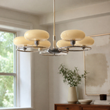 A modern six-light chandelier with cream-colored oval glass shades and a polished silver metal frame, hanging in a bright, contemporary room with a window, console table, and framed artwork.