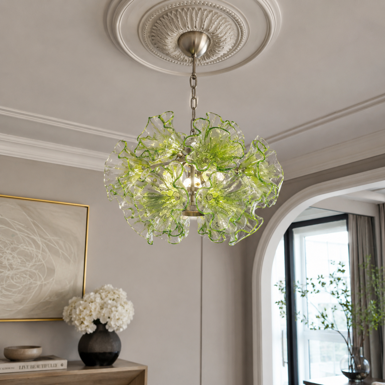 A modern glass chandelier with a ruffled, flower-like silhouette made of clear and light green glass, hanging from a silver chain in a decorated interior room.