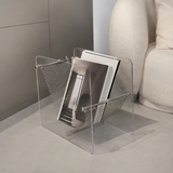 Clear acrylic and metal magazine holder with books and magazines, styled in a minimalist modern room next to a cream-colored armchair.