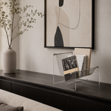 A clear acrylic and metal wire magazine rack holding books and a magazine, styled on a dark wooden console table in a modern living room setting with a vase and framed wall art.