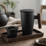 A matte black ceramic tea cup with a wooden handle and lid, shown on a wooden tray alongside a small black tea infuser cup. The scene is styled on a wooden table with a blurred traditional teapot and plants in the background.