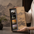 A person holds an open gift box containing four small ceramic cups with mountain-inspired designs in different colors. The box lid features a traditional landscape painting, and the background shows a softly blurred interior setting with a vase of greenery.