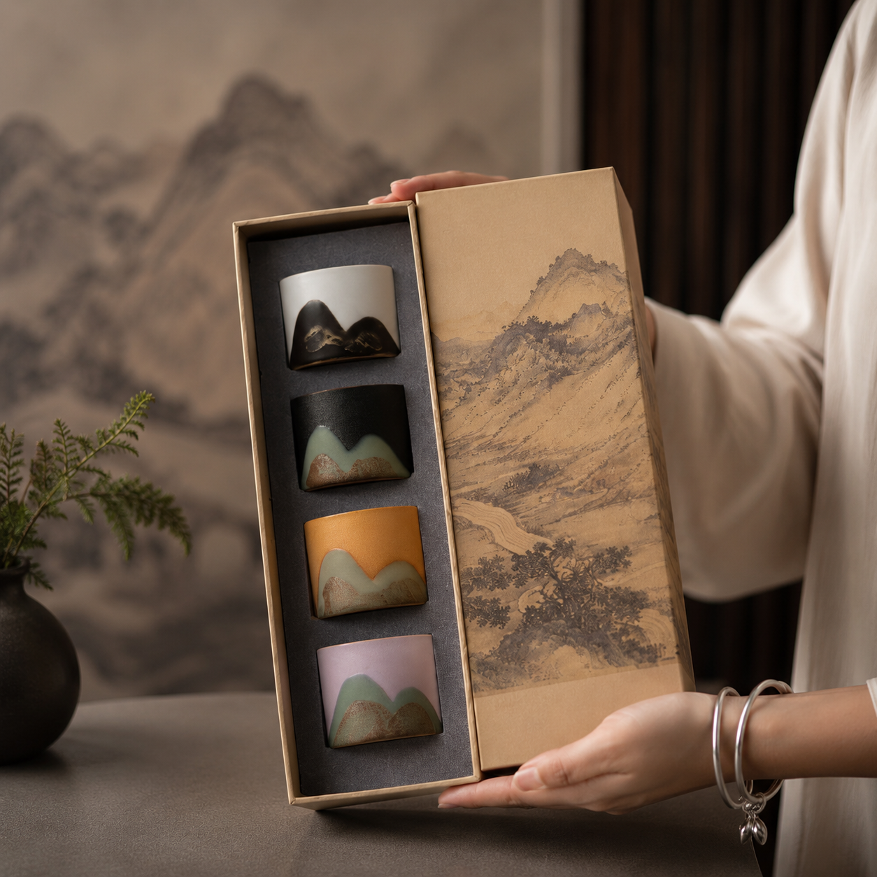 A person holds an open gift box containing four small ceramic cups with mountain-inspired designs in different colors. The box lid features a traditional landscape painting, and the background shows a softly blurred interior setting with a vase of greenery.