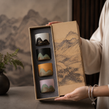 A person holds an open gift box containing four small ceramic cups with mountain-inspired designs in different colors. The box lid features a traditional landscape painting, and the background shows a softly blurred interior setting with a vase of greenery.