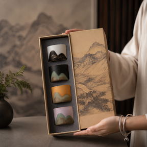 A person holds an open gift box containing four small ceramic cups with mountain-inspired designs in different colors. The box lid features a traditional landscape painting, and the background shows a softly blurred interior setting with a vase of greenery.