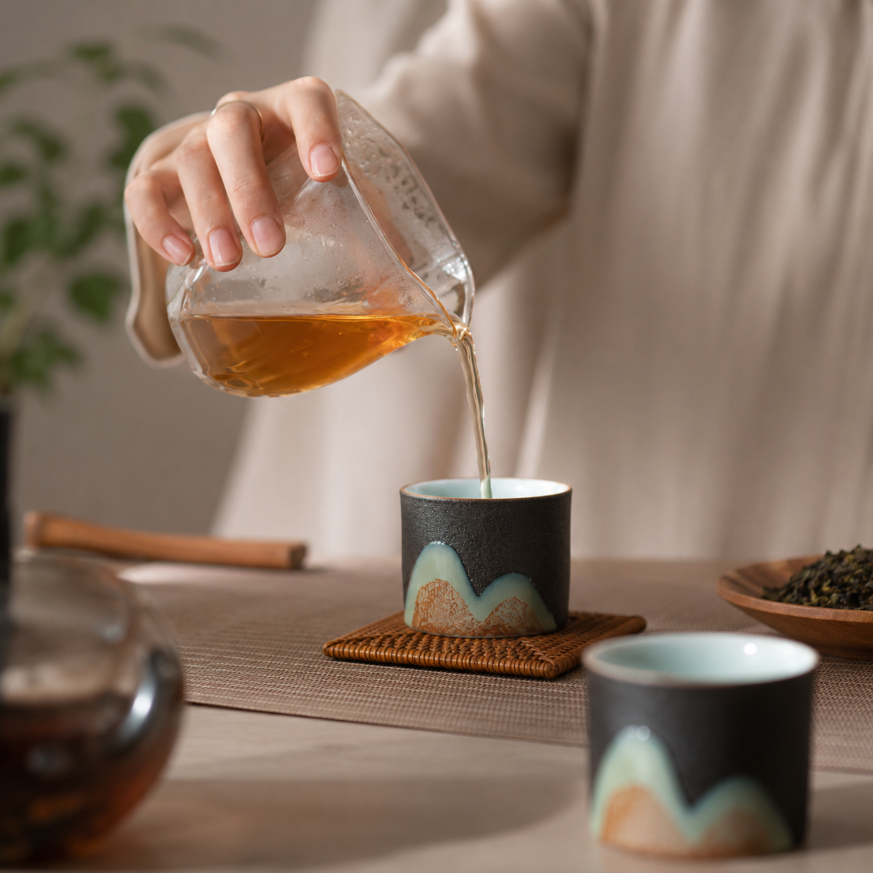 A person in a beige robe pours amber tea from a clear glass pitcher into a small dark ceramic cup with a pale blue and beige wave design. A second matching cup sits in the foreground on a woven mat, with tea accessories softly blurred in the background.