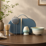 A tea preparation set arranged on a wooden table, including a blue zippered carrying case, a metal strainer, bamboo whisk in a glass holder, a small ceramic vessel, and a bowl with a pale green and cream glaze. A plant and neutral wall are visible in the background.