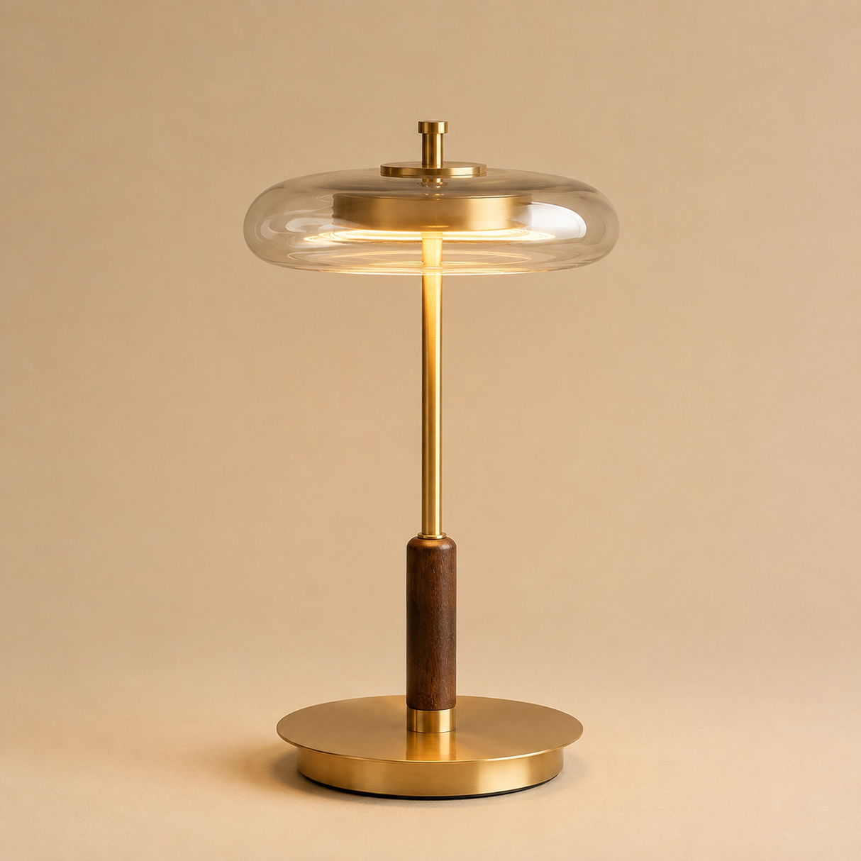 A modern table lamp with a clear glass dome shade, a brushed brass stand, a dark brown wooden handle, and a matching round brass base, set against a solid beige background.