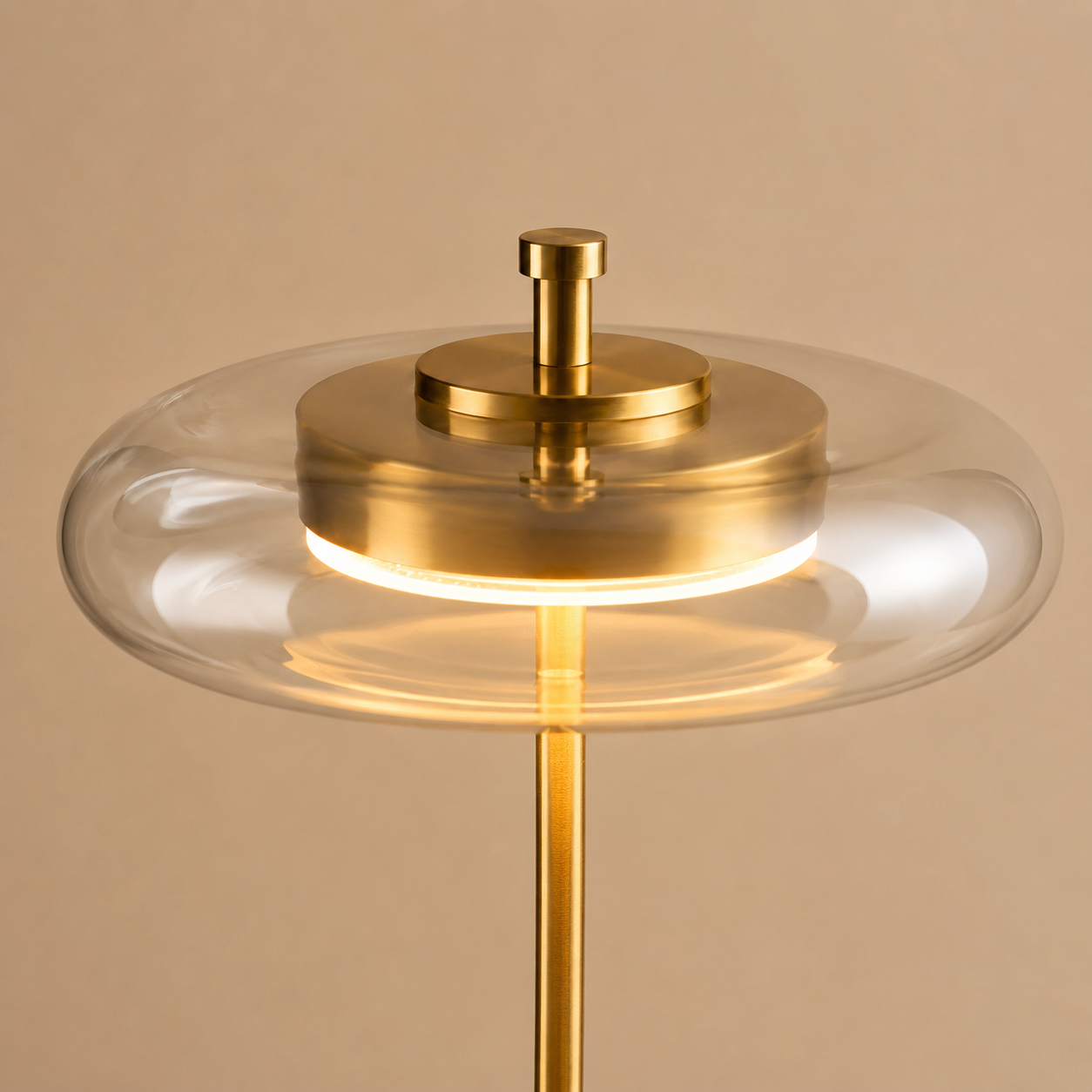 A modern table lamp with a brushed gold brass stand and central cap, featuring a clear glass shade that surrounds a glowing warm white LED light.