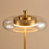 A modern table lamp with a brushed gold brass stand and central cap, featuring a clear glass shade that surrounds a glowing warm white LED light.