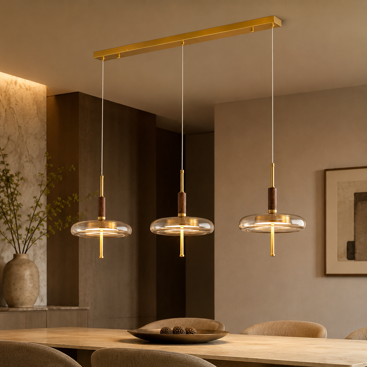 Three-light linear pendant chandelier with a gold-finished metal frame, clear glass shades, and dark brown wood accents, hanging over a dining table in a modern warm-toned interior.