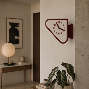 A modern wall clock with a dark red geometric frame and a white grid face, mounted on a beige wall in a styled interior setting with a plant, console table, and lamp.