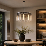 A modern cylindrical pendant chandelier with a textured clear glass shade and vertical hanging rods, illuminated with warm light. It hangs over a round wooden dining table in a styled dining room with a dark vase of greenery and built-in shelving in the background.