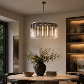A modern cylindrical pendant chandelier with a textured clear glass shade and vertical hanging rods, illuminated with warm light. It hangs over a round wooden dining table in a styled dining room with a dark vase of greenery and built-in shelving in the background.