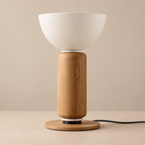 A modern table lamp with a matte white bowl-shaped shade, a cylindrical natural oak wood base, and a round wooden foot ring, shown on a matching beige surface against a neutral beige background.