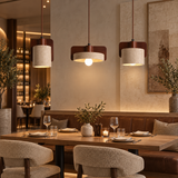 Three modern pendant lights with cylindrical beige fabric shades and dark brown wooden accents hang over a warmly lit dining table in a stylish restaurant setting.