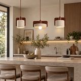 Three modern pendant lights with white drum shades and dark wood side panels hang over a kitchen island in a warm, contemporary kitchen setting.