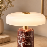 A modern table lamp with a glowing frosted white circular shade and a faceted reddish-brown marble base with white veining, accented by a gold-finished neck and top button.
