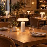 A modern table lamp with a white marble base and a glowing white dome shade sits on a wooden dining table in a warmly lit restaurant setting.