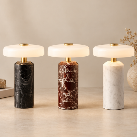 Three modern table lamps with cylindrical marble bases in black, reddish-brown, and white, topped with round white shades and gold accents, arranged on a light beige surface against a neutral background.