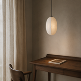 A modern oval pendant light with a warm white, semi-translucent shell hangs over a dark wood desk in a minimalist room with beige curtains and a textured wall.