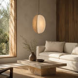 A modern oval pendant light with a warm glowing cream shade hangs over a minimalist beige sofa and stone coffee table in a sunlit, serene living room.