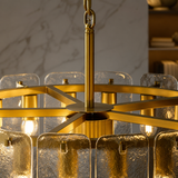 A close-up of a modern chandelier with a brushed gold metal frame, textured clear glass shades, and visible warm light bulbs, shown hanging in an indoor setting.