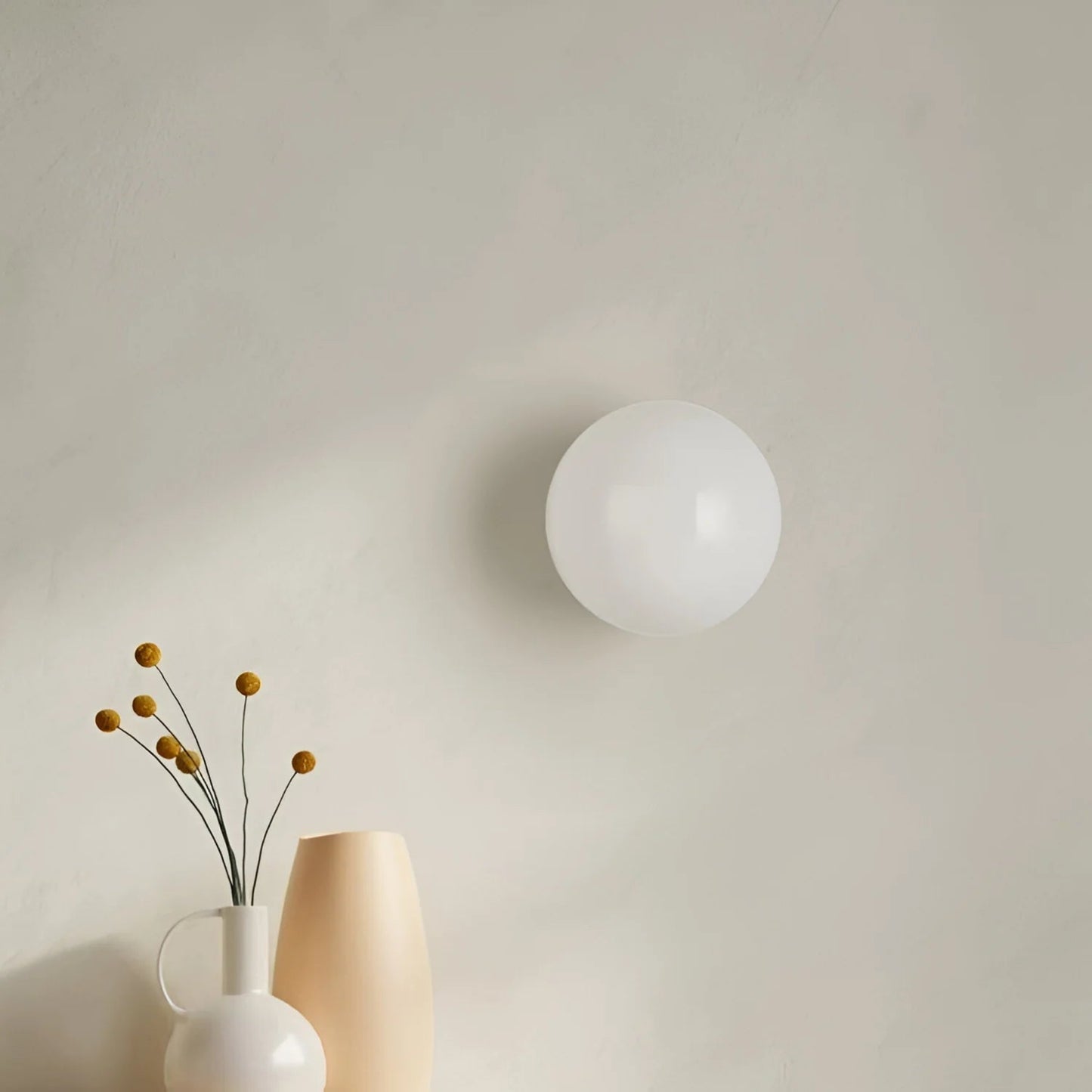 Round wall light fixture on a plain wall with a vase and flowers below.