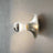 Chrome wall-mounted light fixture on a white wall with a plant in the corner.