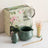 Matcha tea set with green teapot, whisk, and scoop on a white surface with a decorative box in the background.