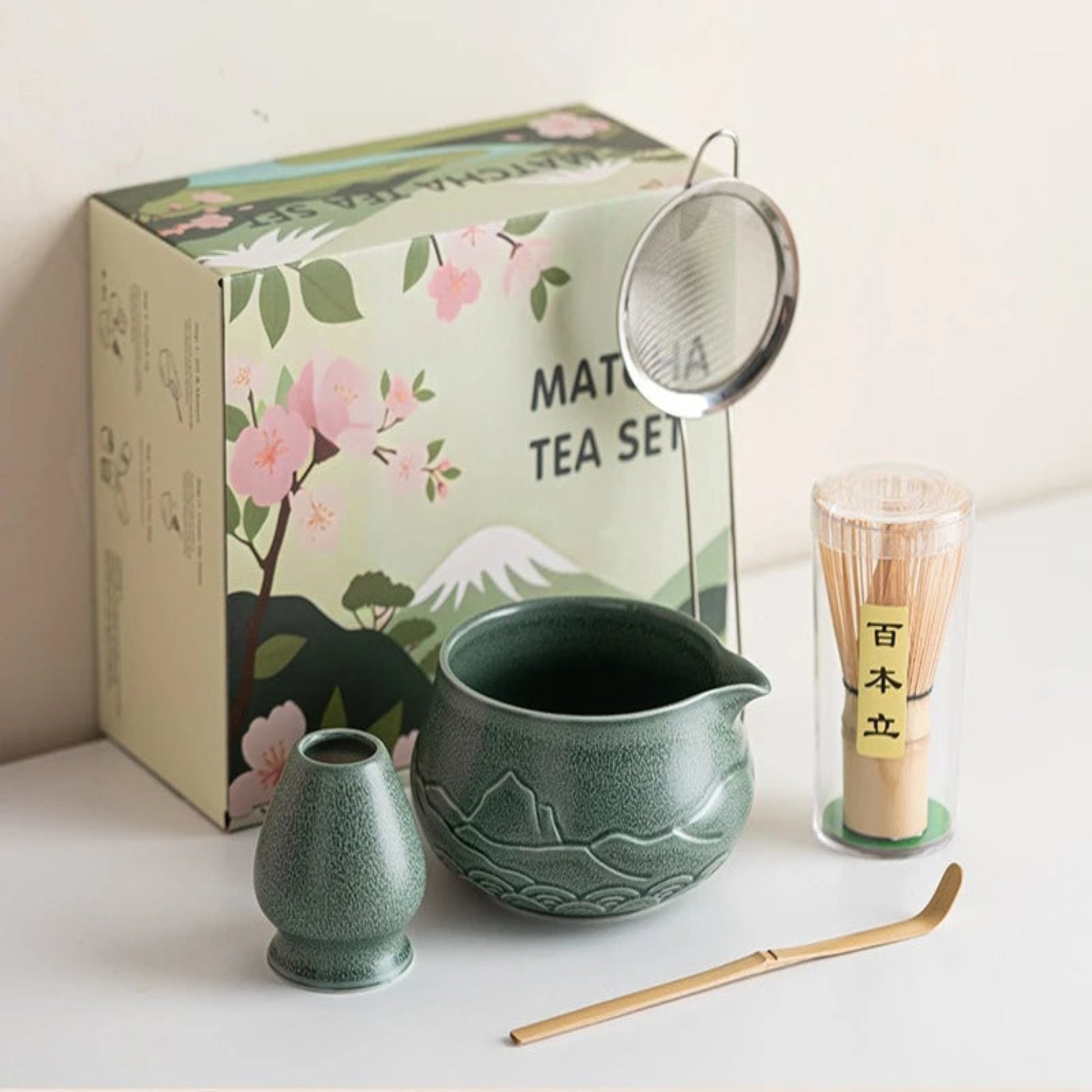 Matcha tea set with green teapot, whisk, and scoop on a white surface with a decorative box in the background.