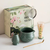Matcha tea set with green teapot, whisk, and scoop on a white surface with a decorative box in the background.