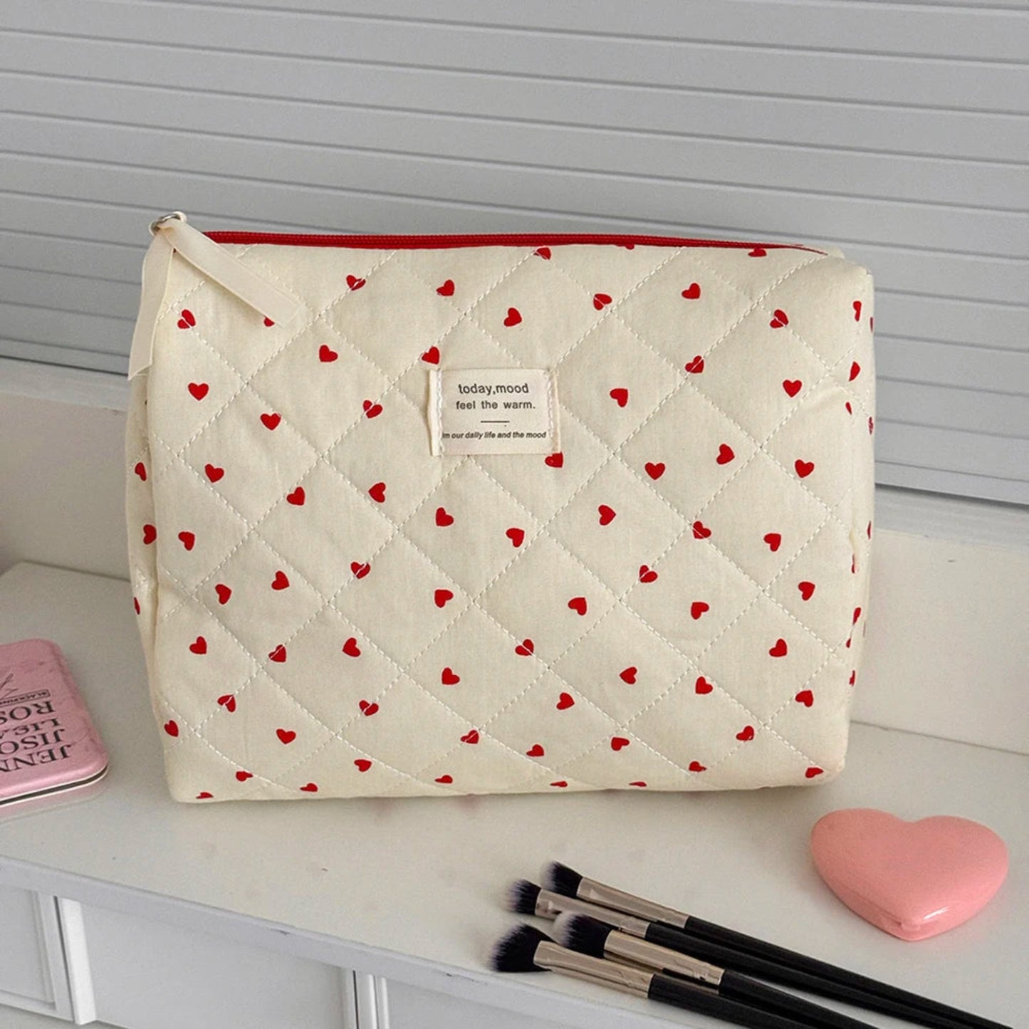 Quilted makeup bag with red heart pattern on a white surface with makeup brushes and a pink heart.