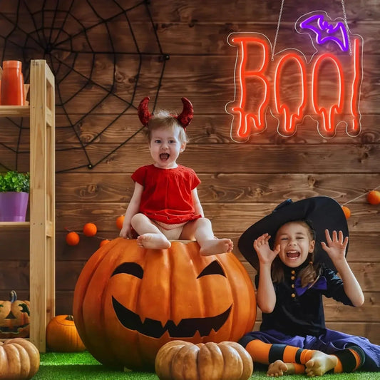 Two children in Halloween costumes sitting on a pumpkin with a 'BOO!' sign in the background.