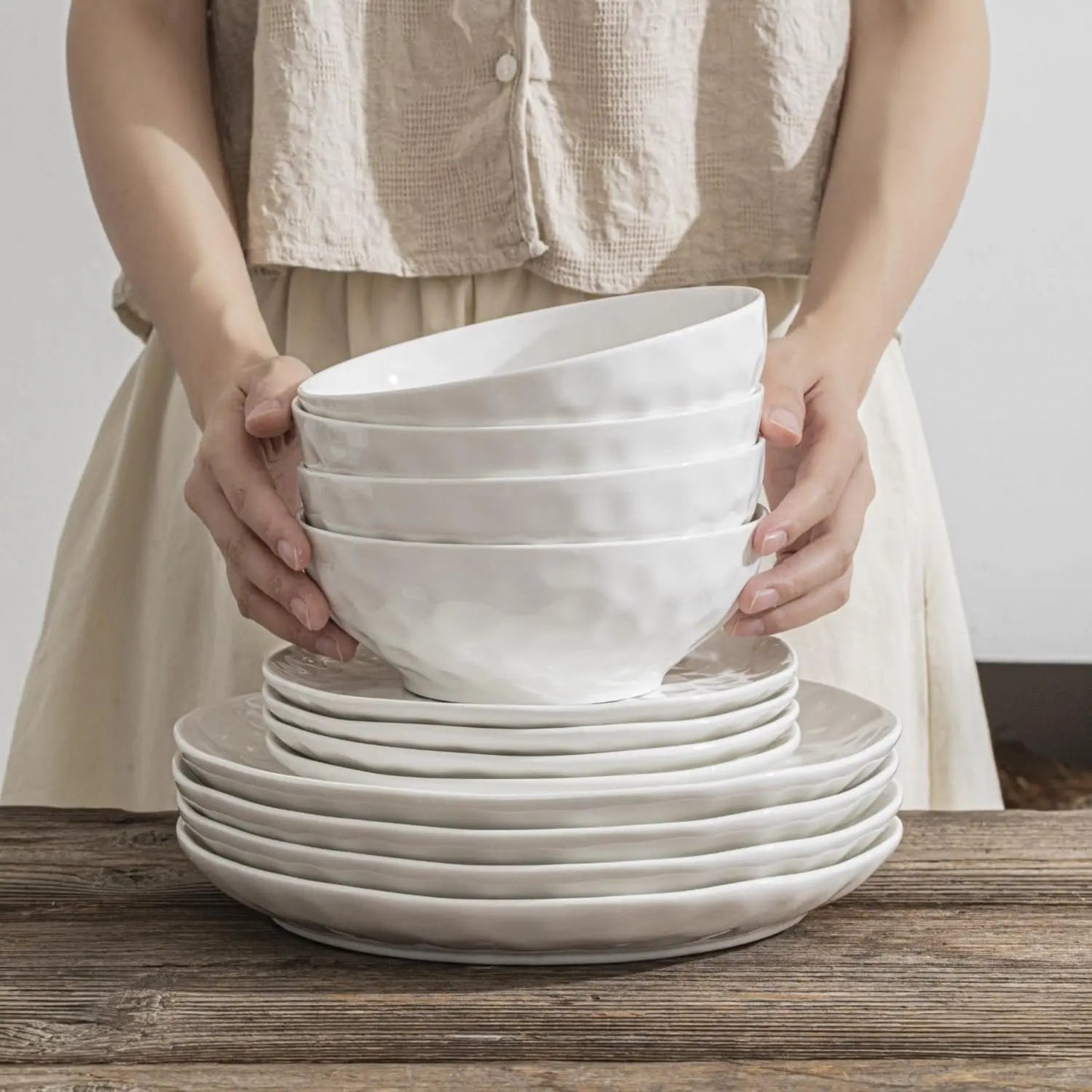 Person holding a stack of white ceramic bowls and plates on a wooden surface.
