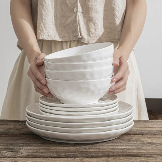 Person holding a stack of white ceramic bowls and plates on a wooden surface.
