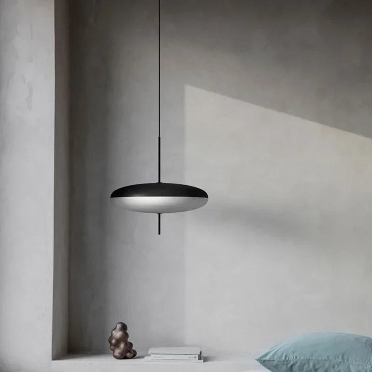 Modern black pendant light hanging in a minimalistic room with concrete walls and a blue pillow.