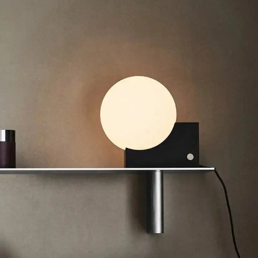 Contemporary LED desk light with dimmable glass ball and matte black base on a shelf. Adds a warm glow to modern interiors.
