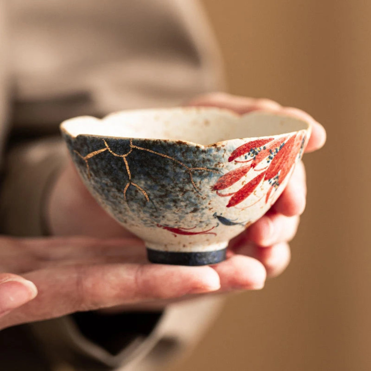 A handmade ceramic bowl with a hand-pan edge, decorated with red fish motifs and gold crackle mikiashi lines on a speckled off-white glaze, held gently in two hands against a warm, blurred background.