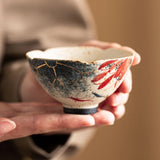 A handmade ceramic bowl with a hand-pan edge, decorated with red fish motifs and gold crackle mikiashi lines on a speckled off-white glaze, held gently in two hands against a warm, blurred background.