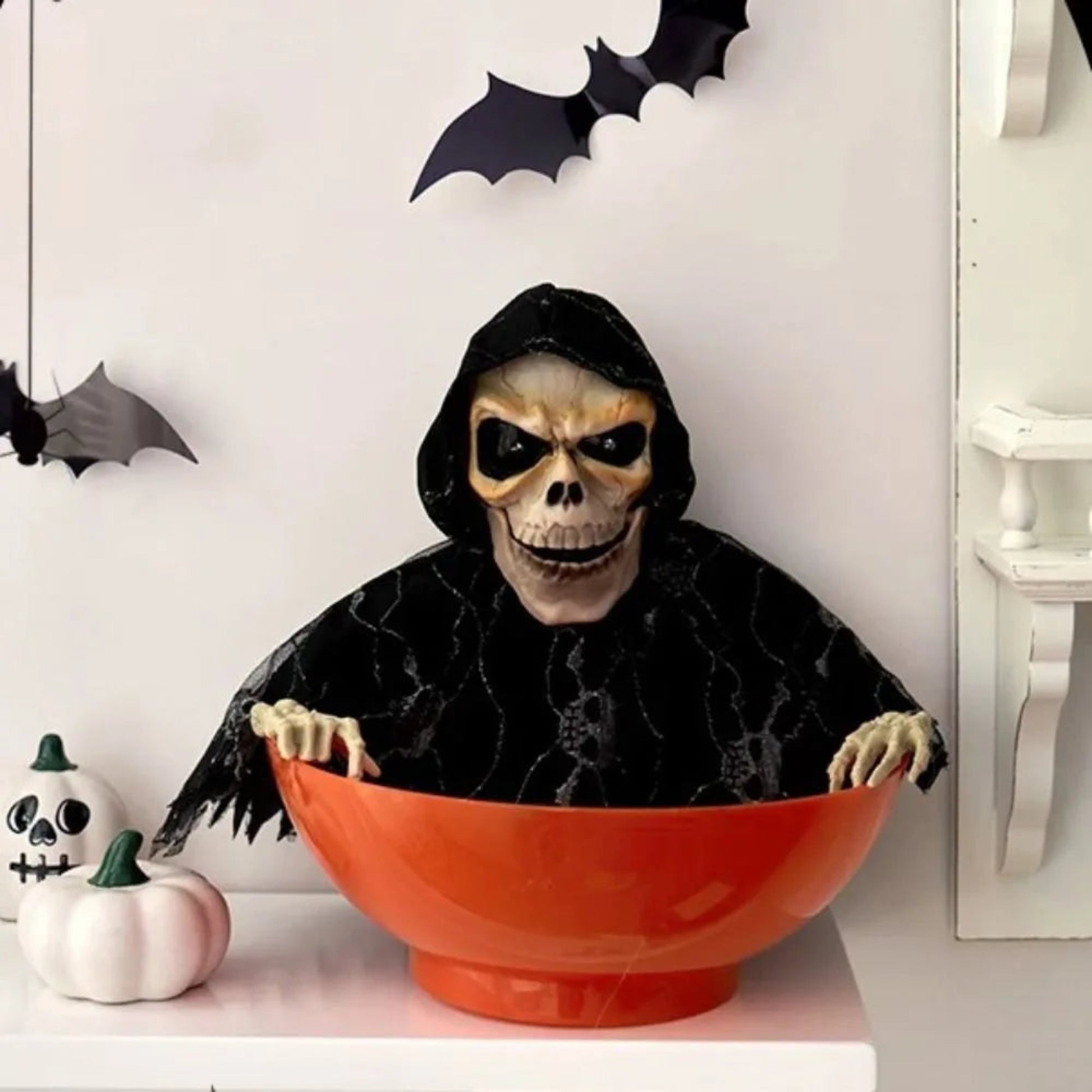 Person in a Halloween costume with a skull mask and black cape sitting inside an orange bowl, with decorative bats and pumpkins in the background.