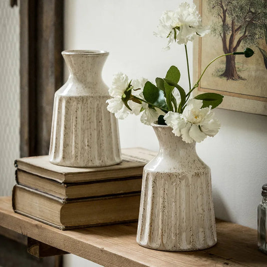 Two ceramic vases on books with a vase of white flowers against a neutral wall.